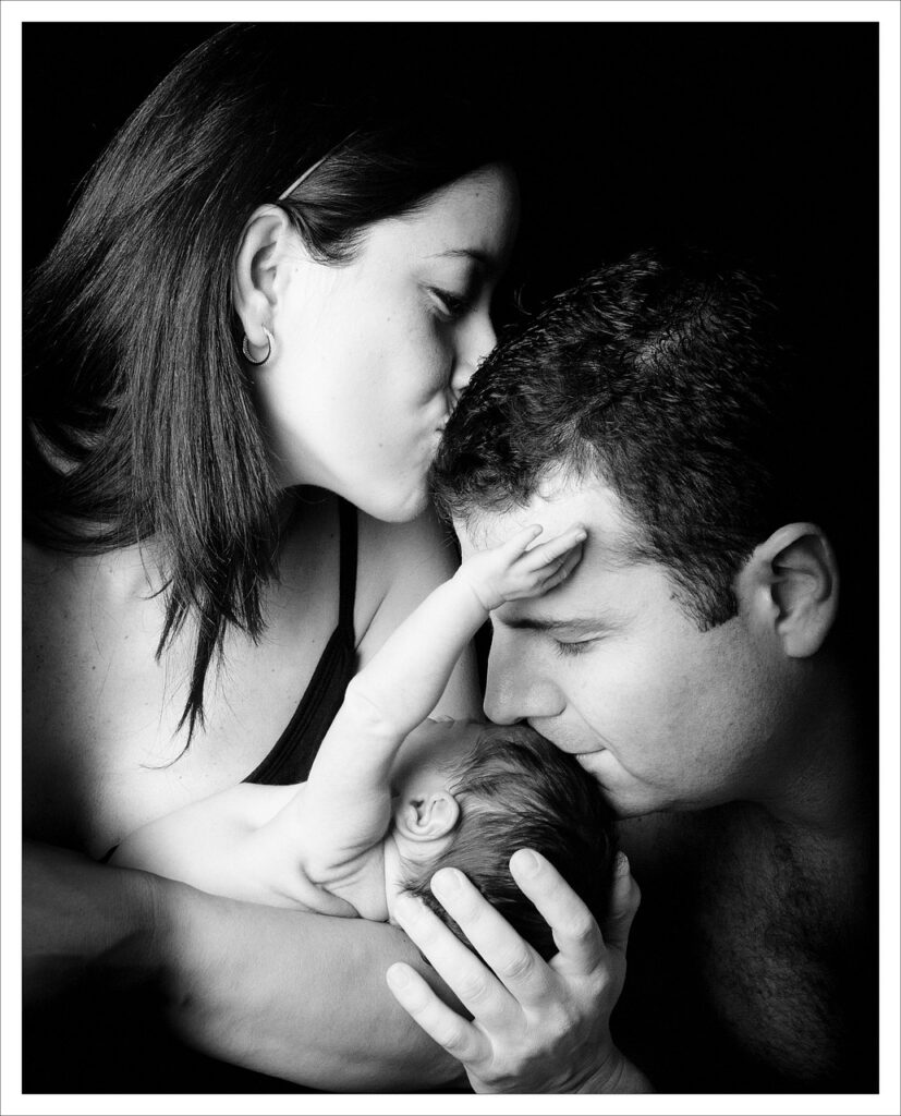 photo of a woman kissing her husbands head as he kisses the head of their baby